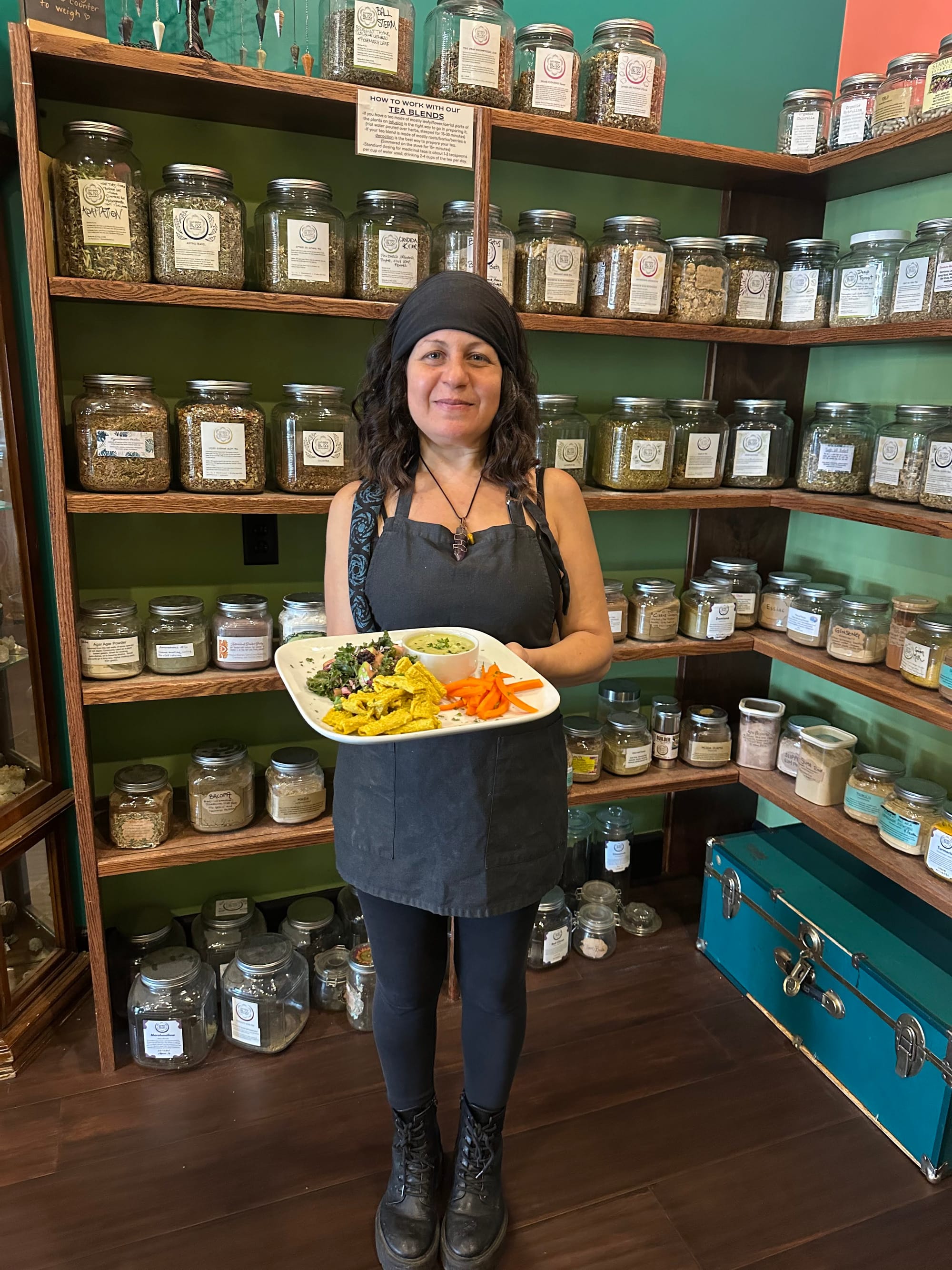 A person in an apron holds a plate in a store with glass jars on shelves. Rainbow Bliss Botanicals is appropriate here.