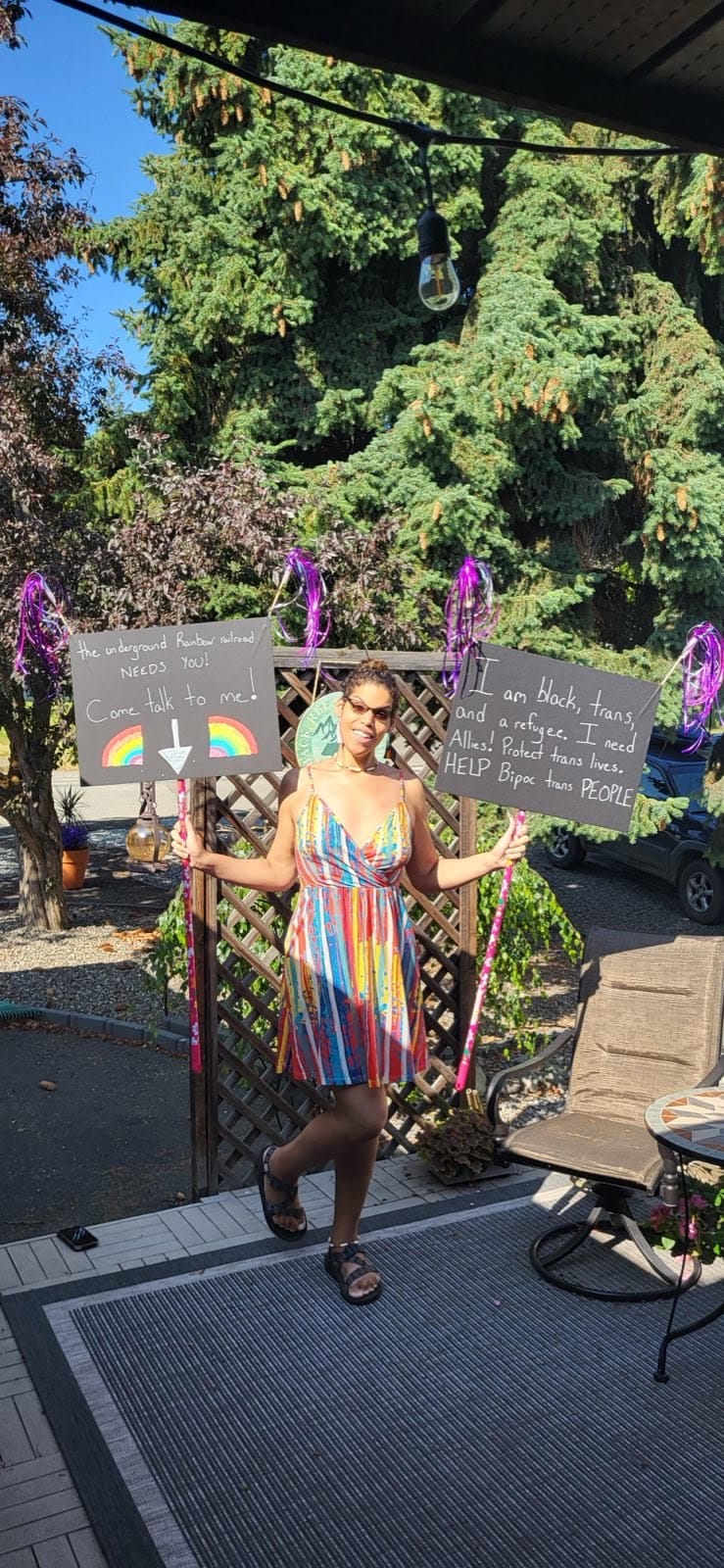 An image of a person in a rainbow dress holding two signs indicating that they are a black, trans refugee.