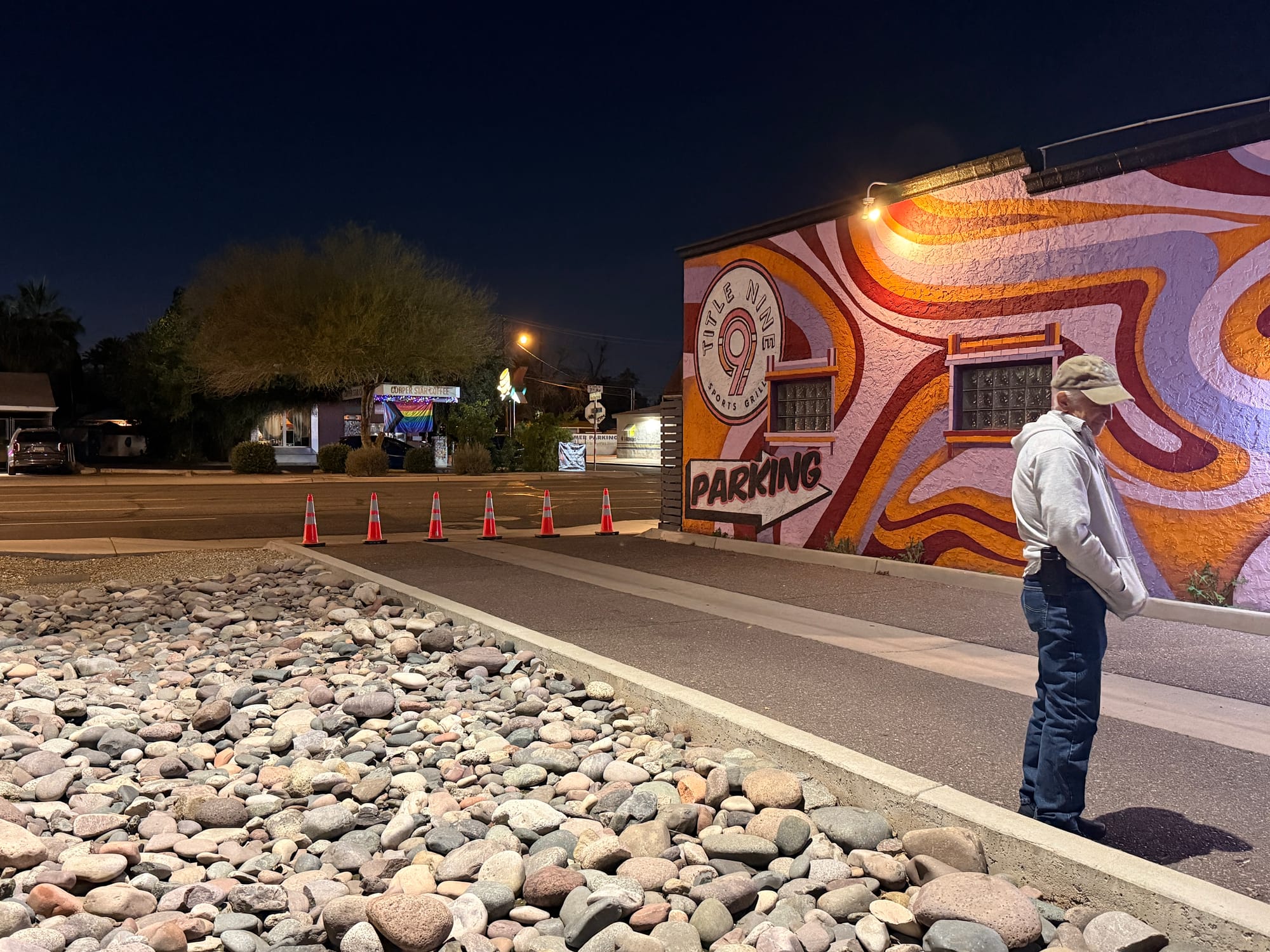 Man standing outside a brightly painted building with a mural at night, orange traffic cones lining the sidewalk.