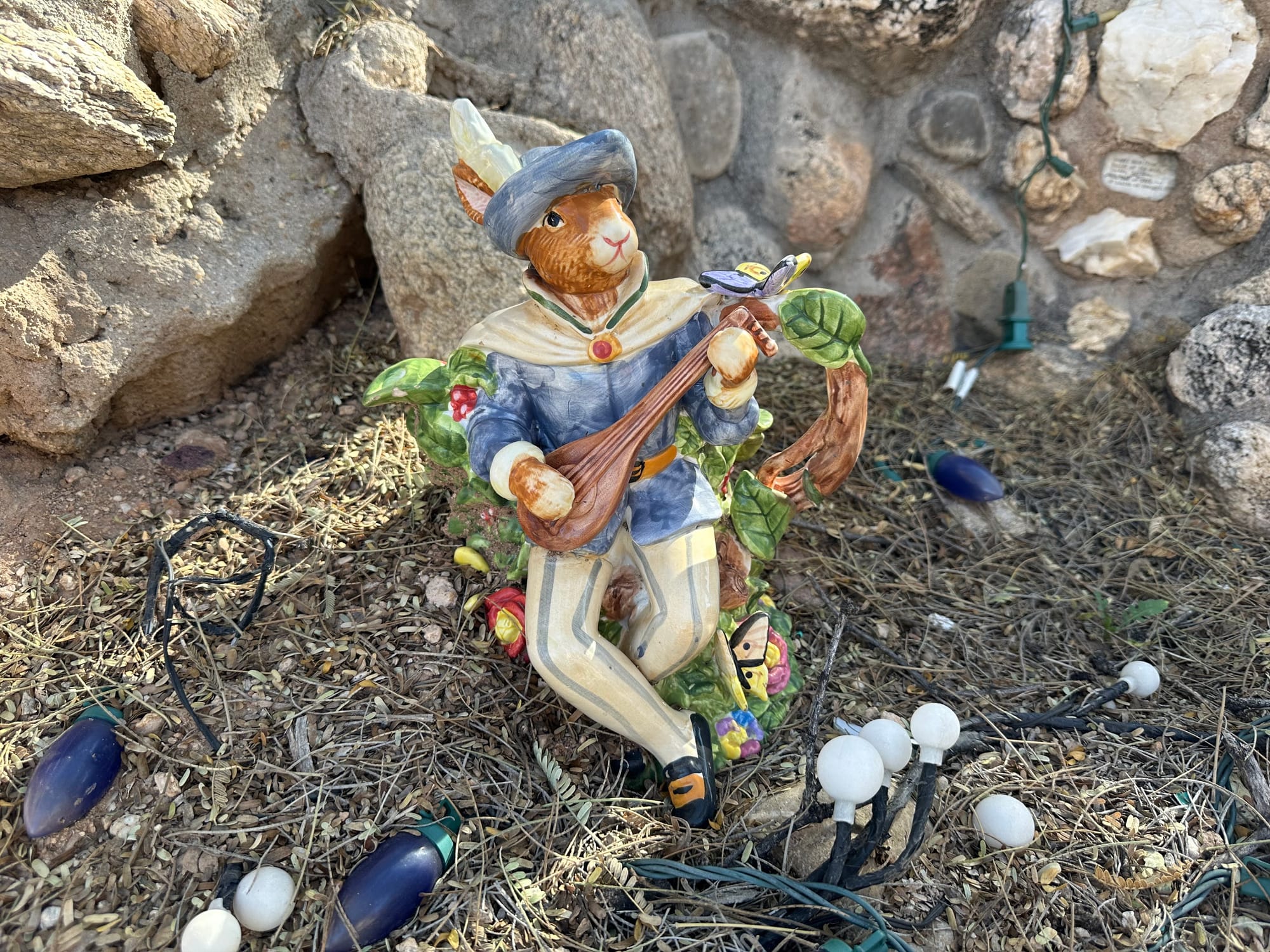 A whimsical cat figurine dressed in medieval attire, playing a stringed instrument, surrounded by lights and rocks.
