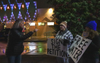 People protesting outside holding signs saying "sign the petition" and "free water" as another person defensively points their finger at them. 