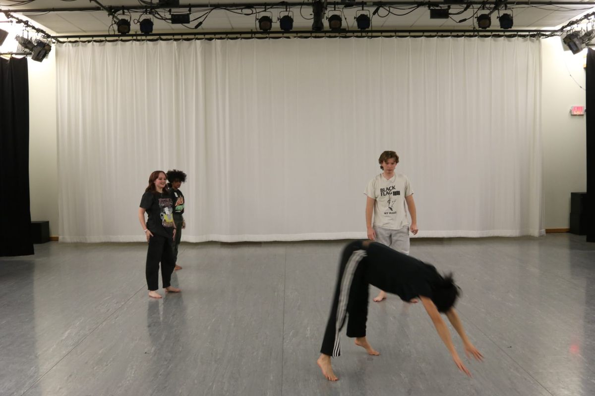 four dancers in front of a white curtain. one in front is leaping to the ground. 