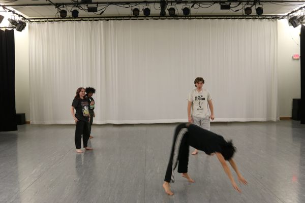 four dancers in front of a white curtain. one in front is leaping to the ground. 