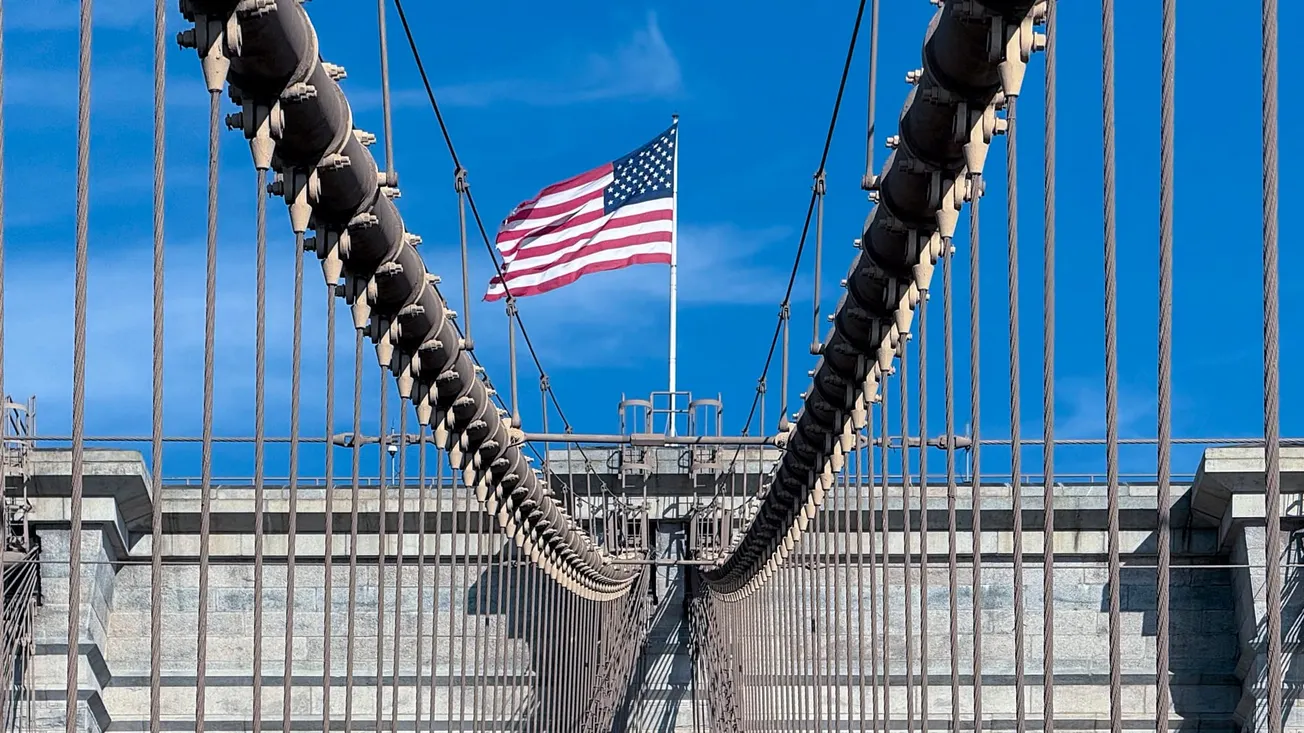 Photo of an American flag flying over the Brooklyn Bridge against a bright blue sky. Taken by Frederick Lane in September 2024.