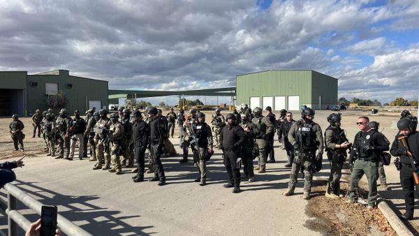 Photo of rural yard with green shops in background and about 50 heavily armed federal agents in the foreground, under dramatic clouds. 