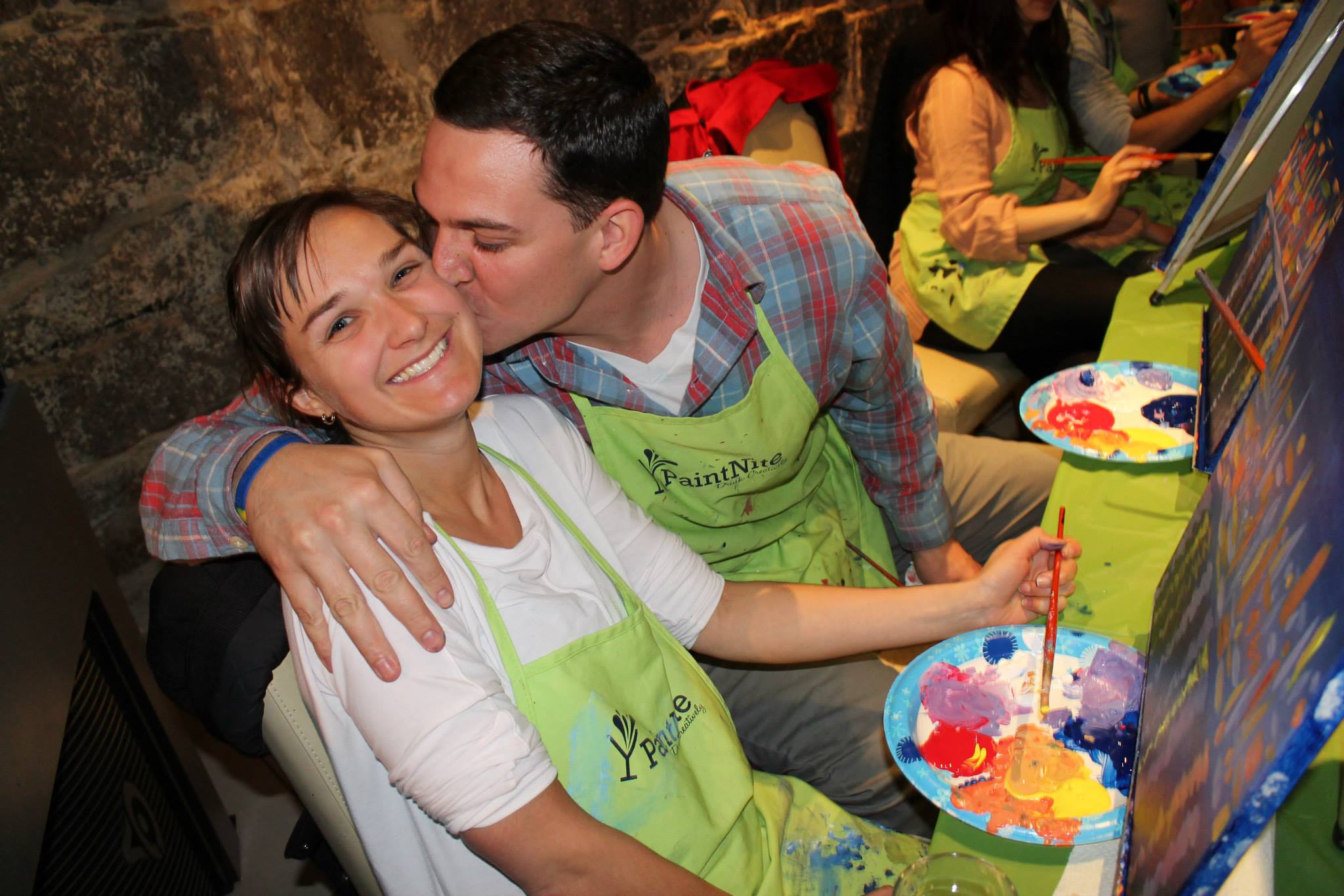 Happy couple at a paint night event in a local bar, embracing while surrounded by other couples painting