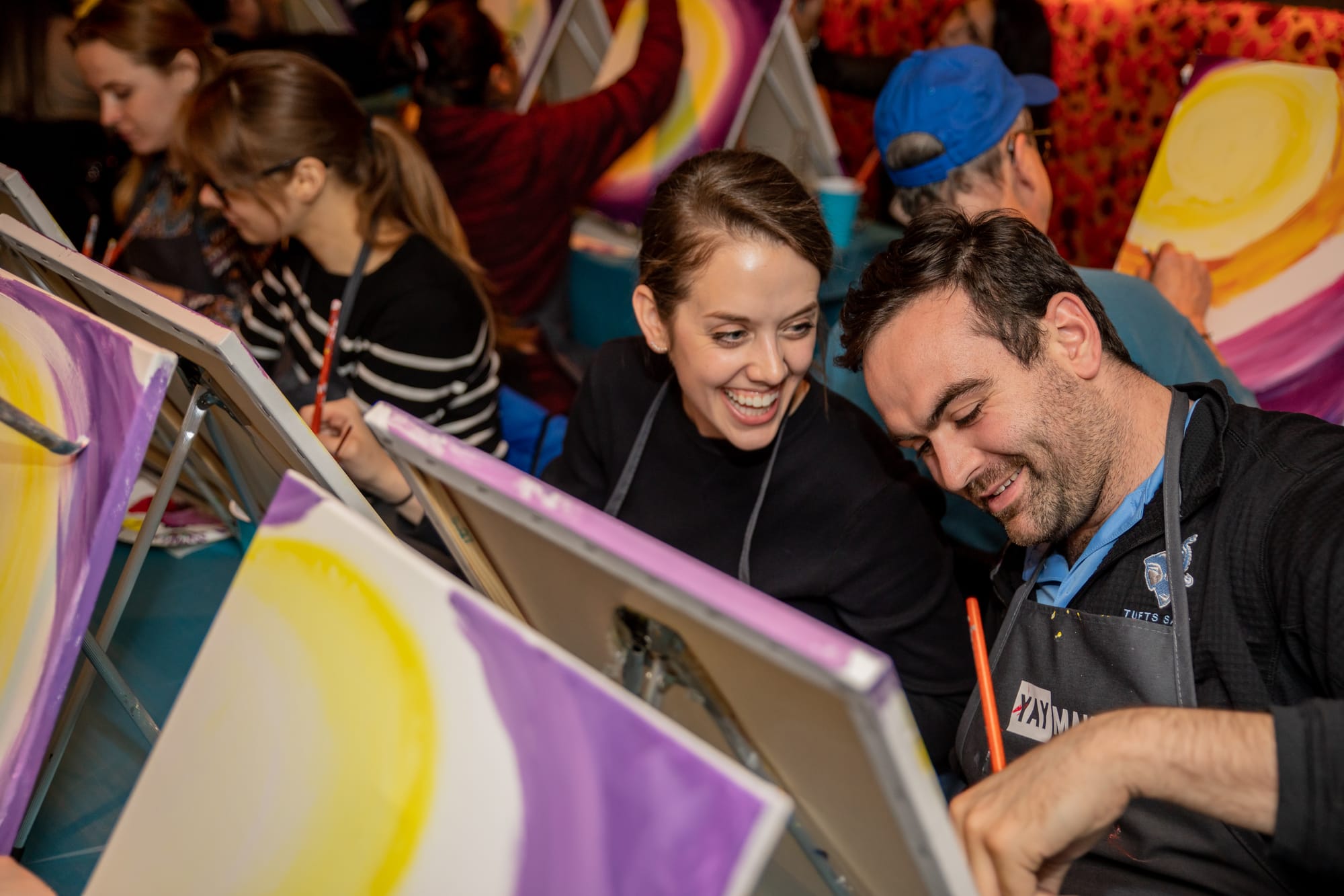 Couple laughing during paint and sip date night at a bar venue, holding paintbrushes while creating art together