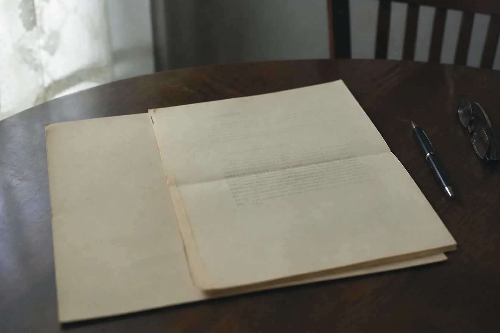 A yellowed mortgage note lying open on a dark wooden table, its pages creased from folding, with a pen and reading glasses beside it.