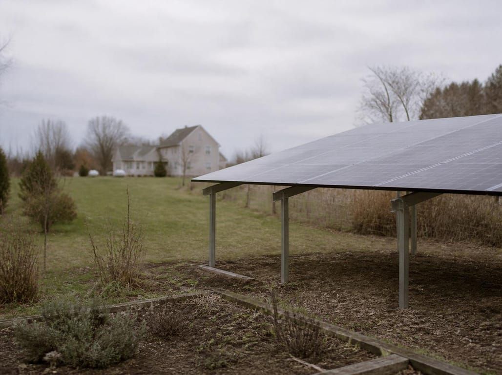 A ground-mounted solar array on steel posts standing at the far end of a garden, with the house some distance behind it on an overcast day.