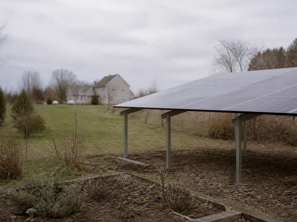 A ground-mounted solar array on steel posts standing at the far end of a garden, with the house some distance behind it on an overcast day.