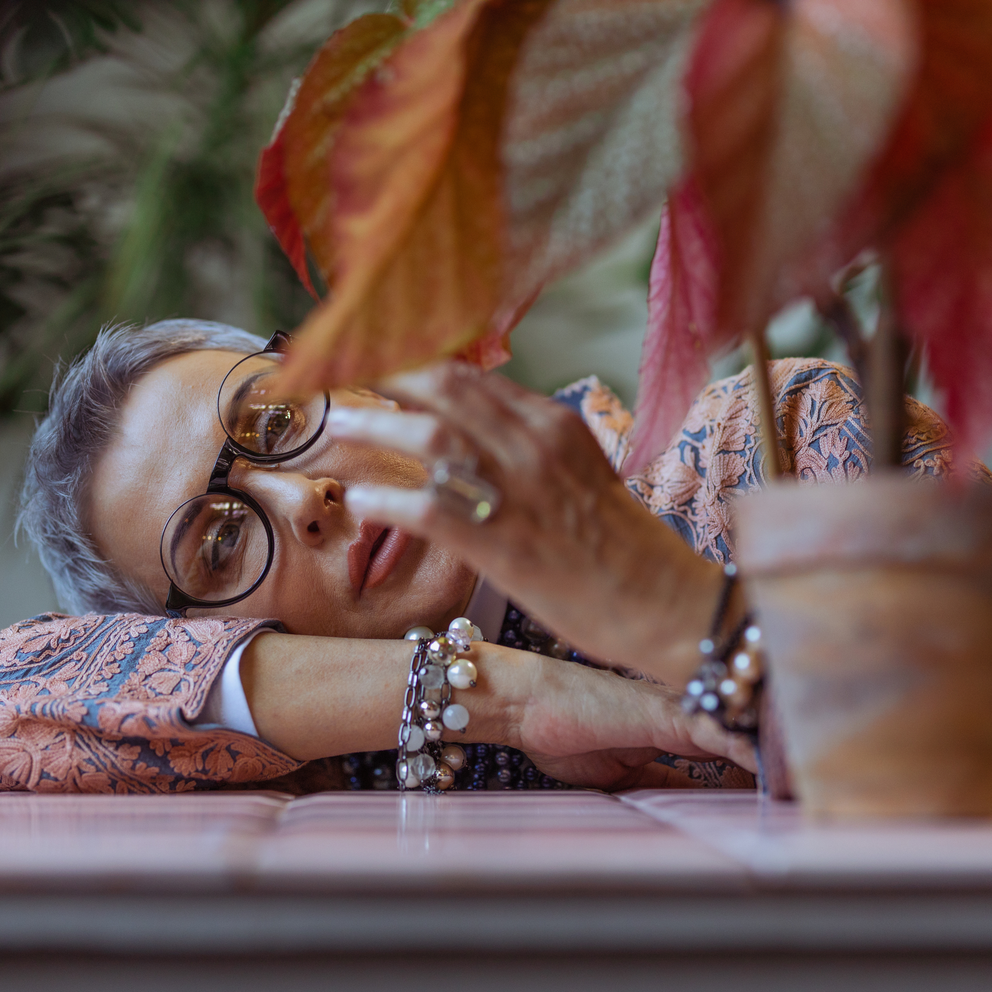 photo of a middle age woman resting her head on a counter and touching the leaves of a potted plant.