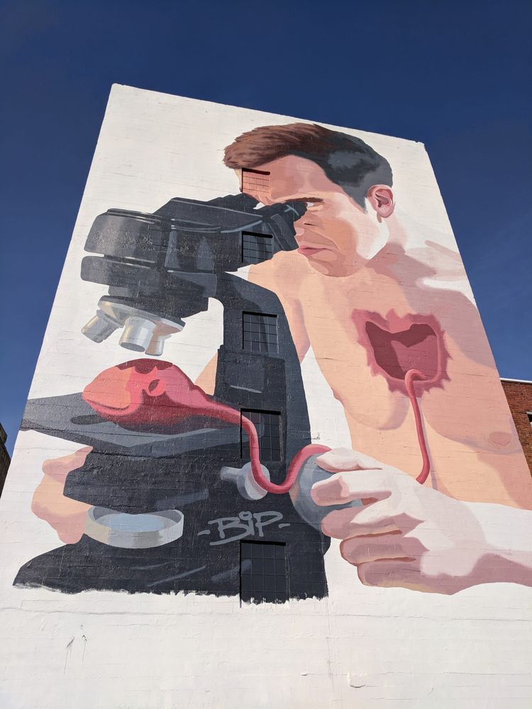 A large mural painted on the side of a building shows a man examining his own heart on a microscope, the heart is still attached to his body