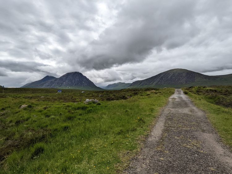 A gravel path runs through green moorland, dotted with small yellow wildflowers, toward a range of dark mountains. A steep, pointed peak rises on the left with its summit hidden in cloud, and