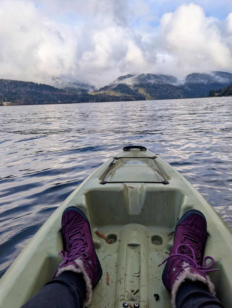 a photo shows the end of a kayak and two purple snowboots on a lake facing a coastline of rolling hills with snow on them
