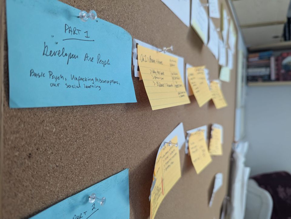 a corkboard has blue and yellow postit notes on it with handwritten notes. The blue postit note in focus reads Part 1: Developers are People.