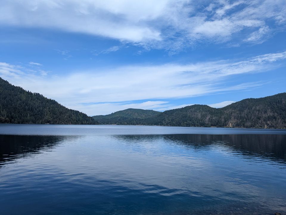 A photo of a lake with clear water. There are hills behind it covered in pine trees, and a blue sky with some streaks of cloud