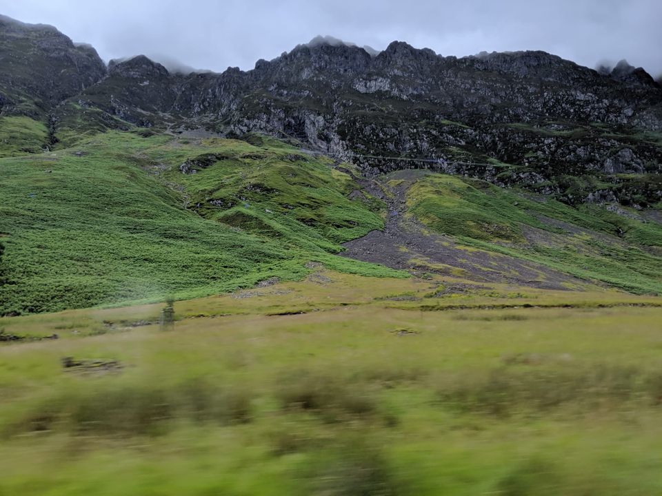 A photo shows a green landscape with rocky hills under an atmospheric grey sky with mist. There is motion blur at the bottom suggesting this was taken from a moving vehicle