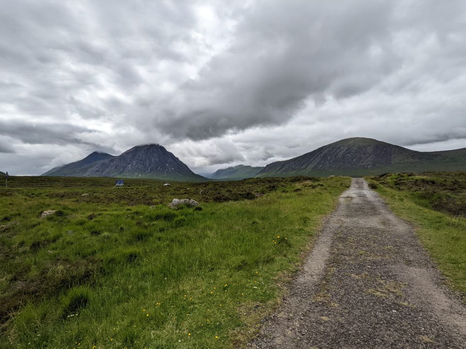 A gravel path runs through green moorland, dotted with small yellow wildflowers, toward a range of dark mountains. A steep, pointed peak rises on the left with its summit hidden in cloud, and