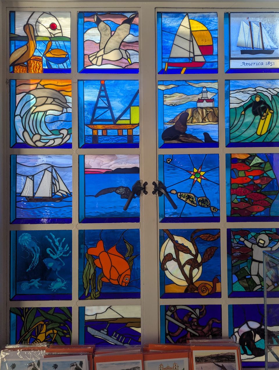 A stained glass window with many small squares depicting ocean scenes such as fish, a surfer, a boat, jellyfish, pelicans and dolphins in a variety of colors with a rich blue backgroundn