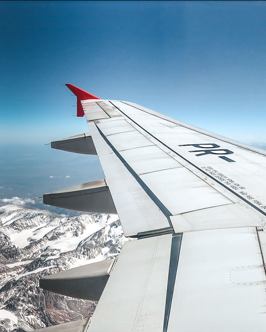 Airplane wing over the Andes Mountains, en route to Chile, with snow-capped peaks below and a clear blue sky.