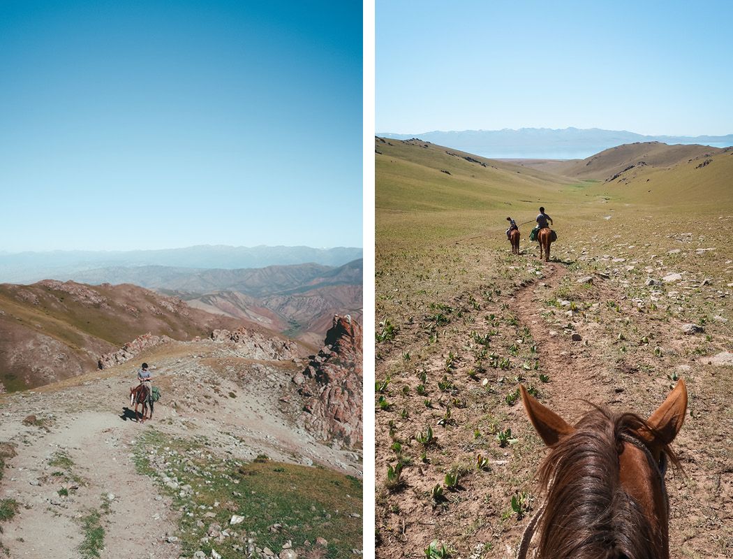 Two horseback riders on a dirt trail through wide, grassy valleys surrounded by rolling hills under a clear blue sky.