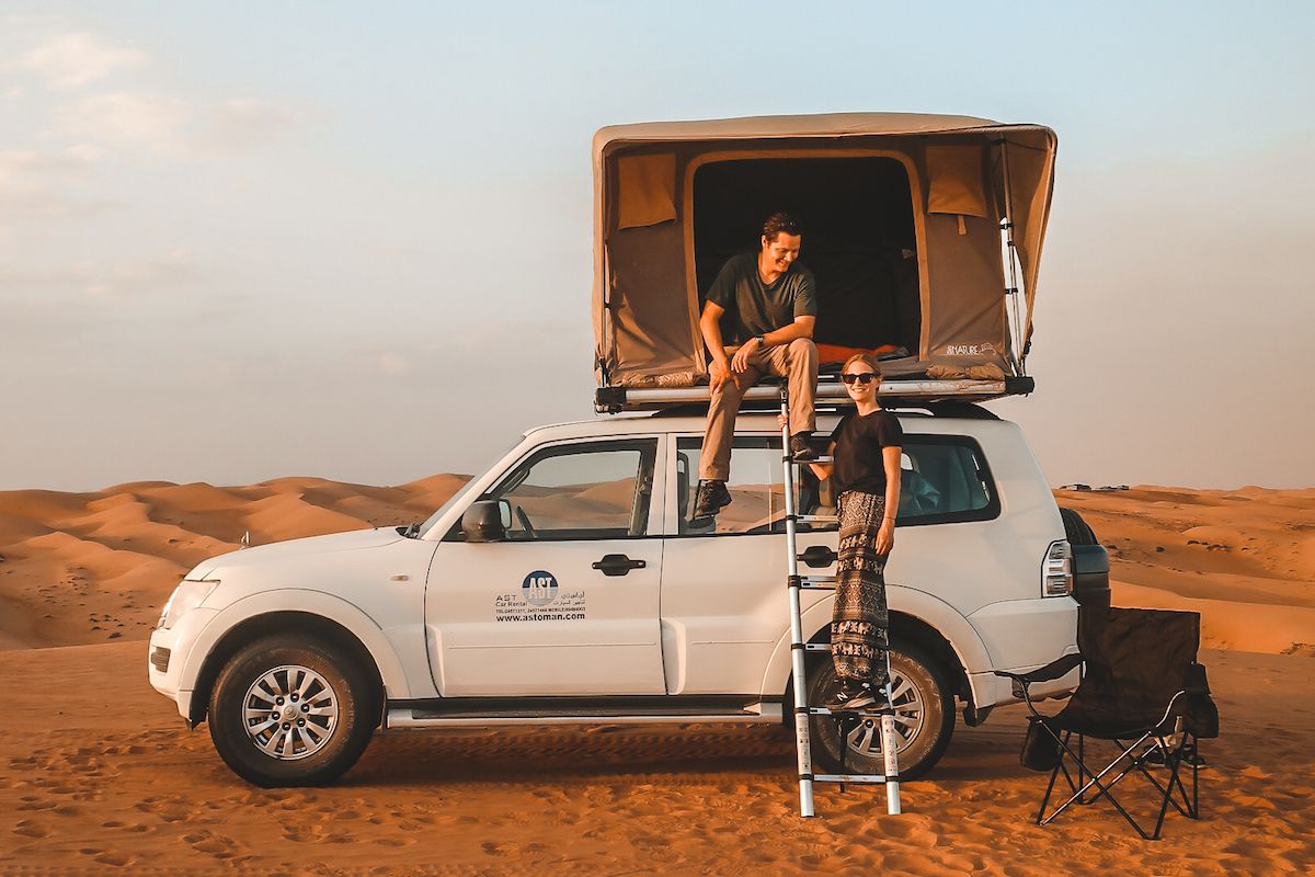 Cec and Ari camps on top of their 4x4 vehicle in the Omani desert, rooftop tent open against a soft sunset glow over sand dunes.