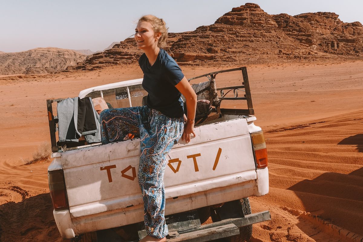 Cec climbs out of the back of a pickup truck during a desert safari in Wadi Rum, Jordan, with red cliffs behind.