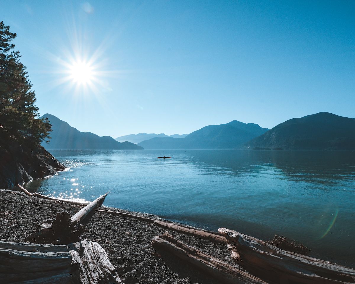 Kayaker glides across calm waters at sunrise on BC's coast, surrounded by mountains and driftwood along the serene shoreline.