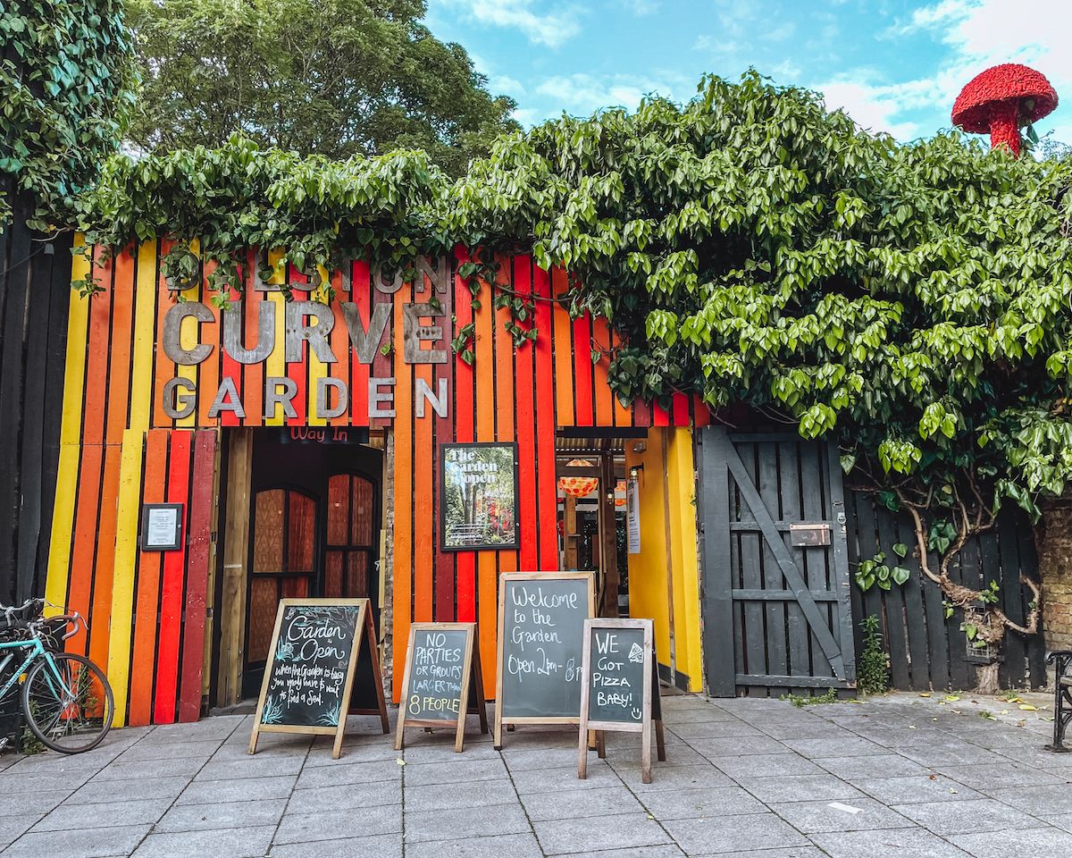 The colourful entrance of Dalston Curve Garden, covered in greenery, with chalkboard signs welcoming visitors and a red mushroom sculpture peeking out.
