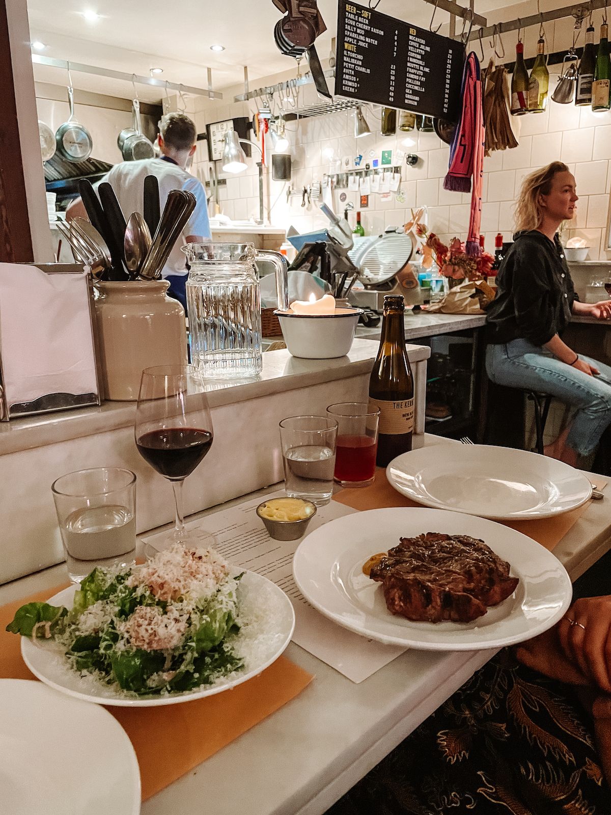 A dinner spread at Hill & Szrok featuring steak and Caesar salad, with an open kitchen in the background.