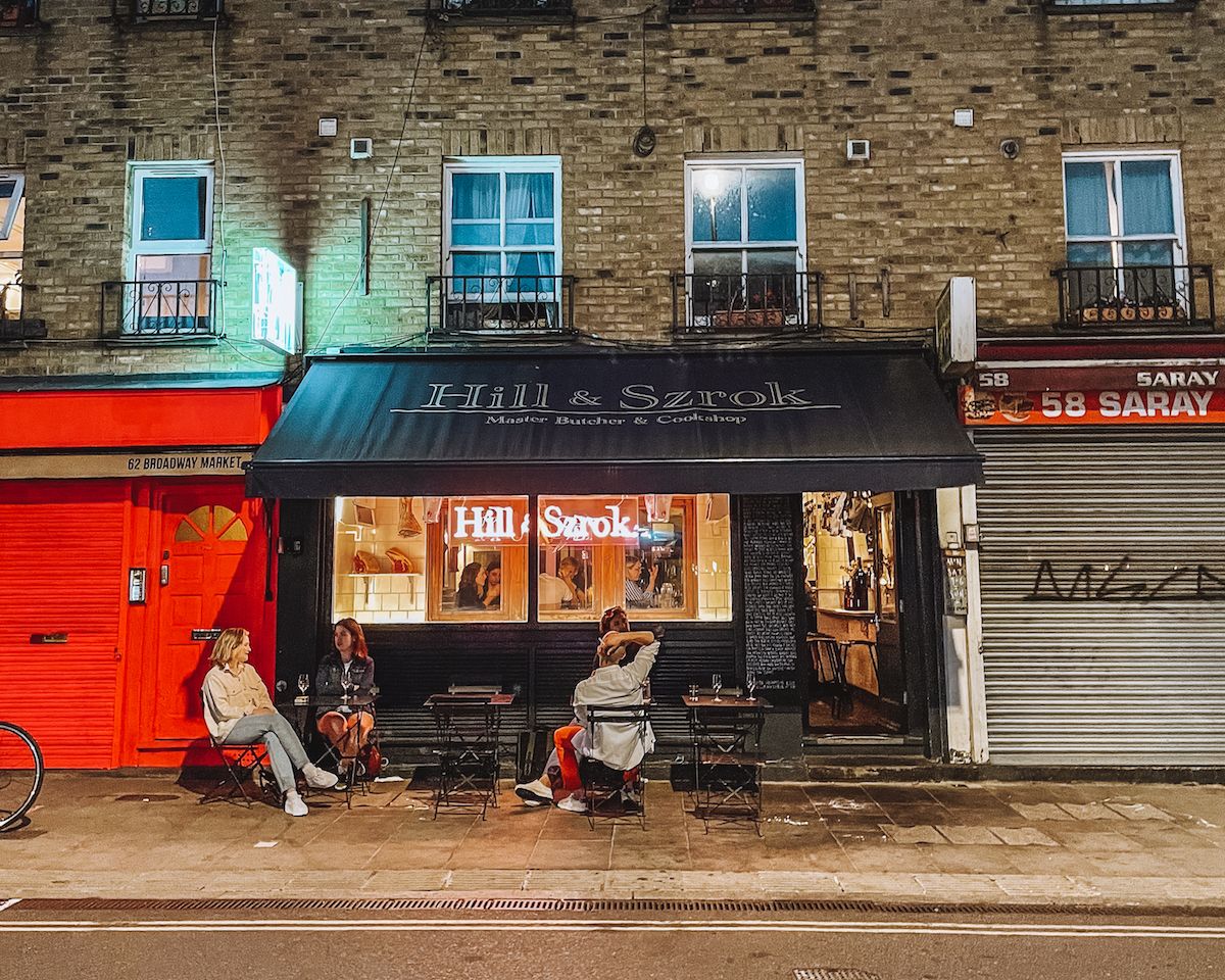 Hill & Szrok butcher and restaurant on Broadway Market at night, with patrons dining outside under its black awning.