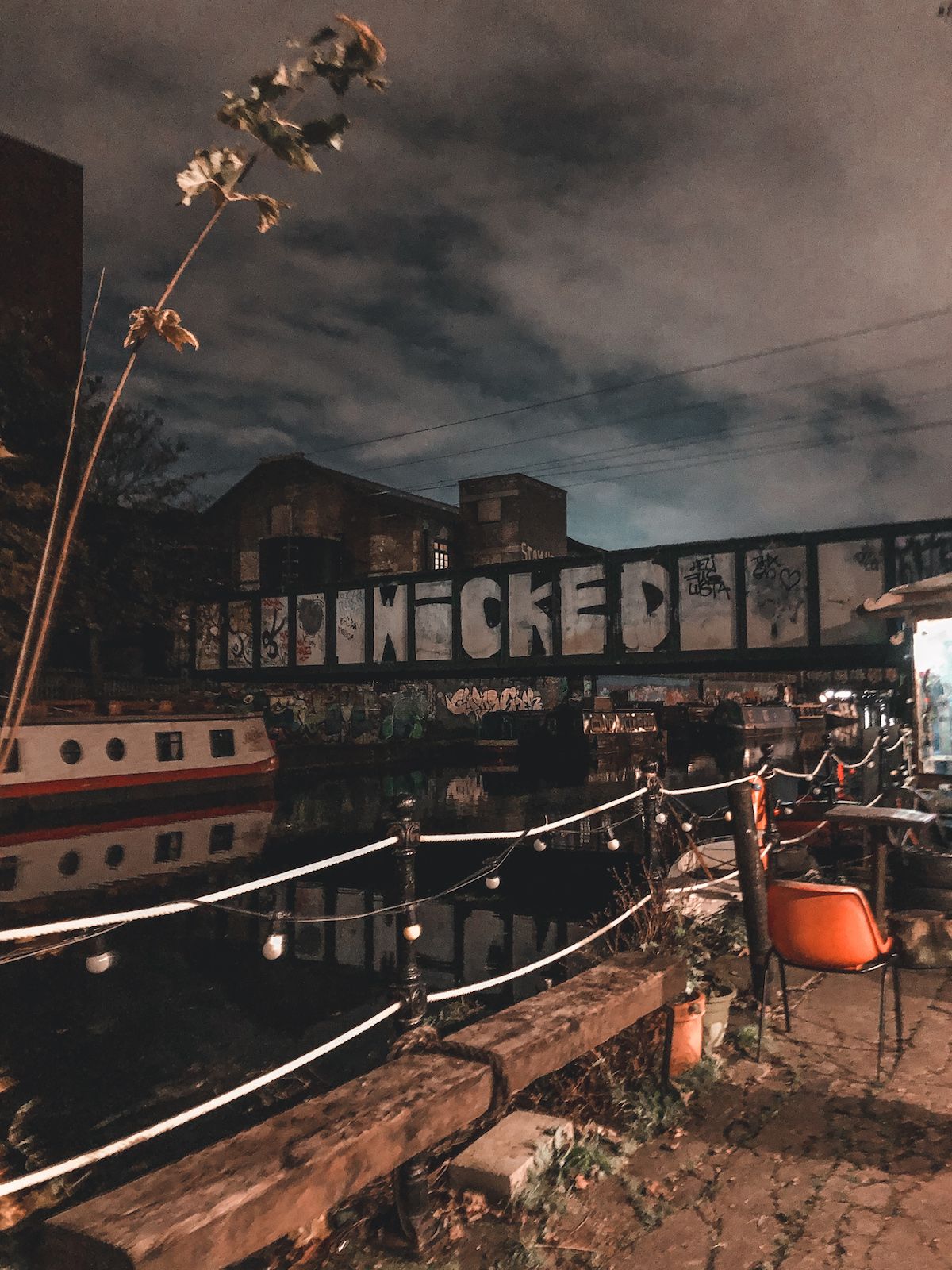 A dimly lit canal in Hackney Wick with a graffiti-covered bridge reading "Wicked," and scattered outdoor seating.