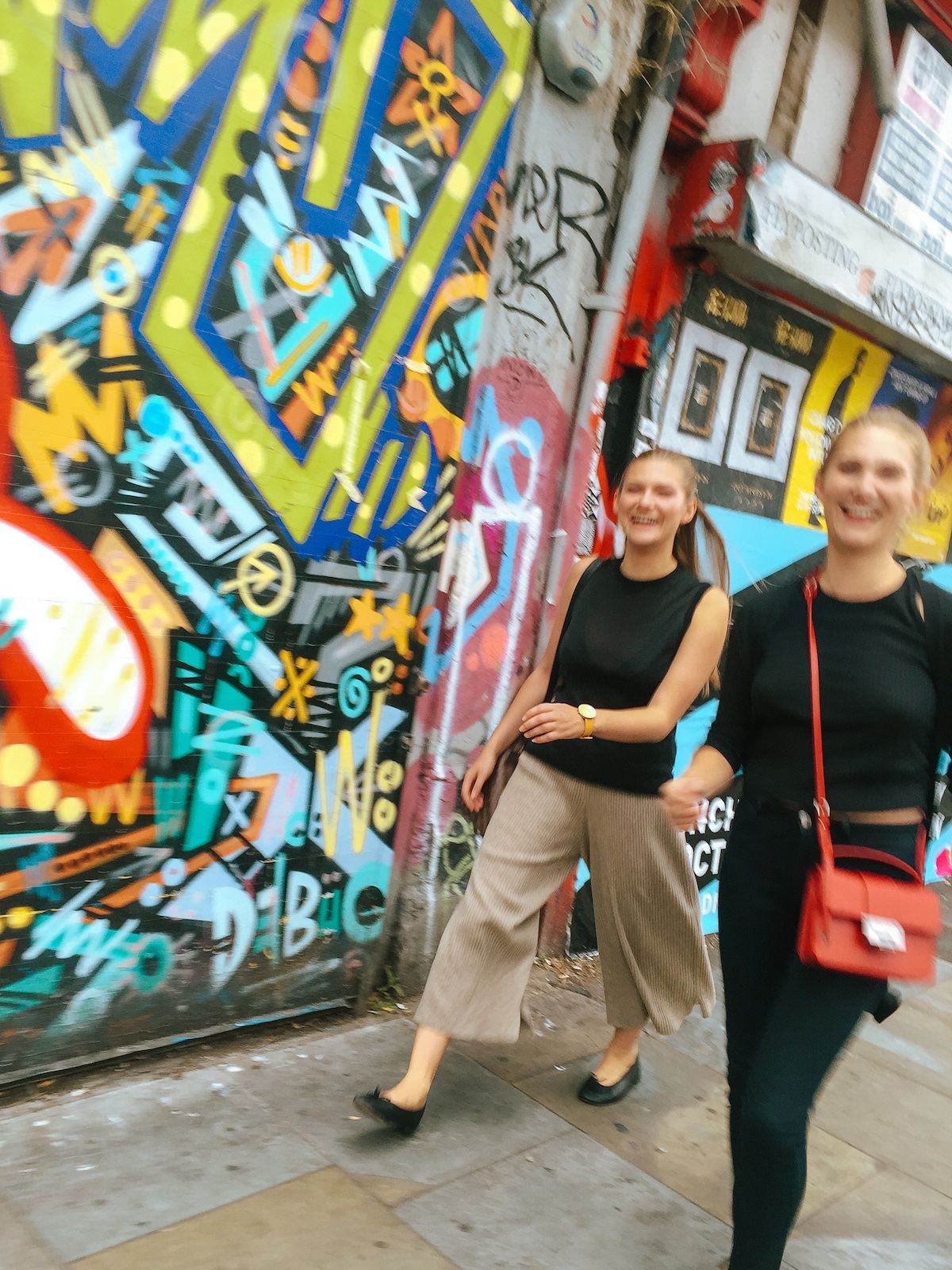 Cec and Katarina walking along walking past a colourful graffiti wall in Shoreditch.