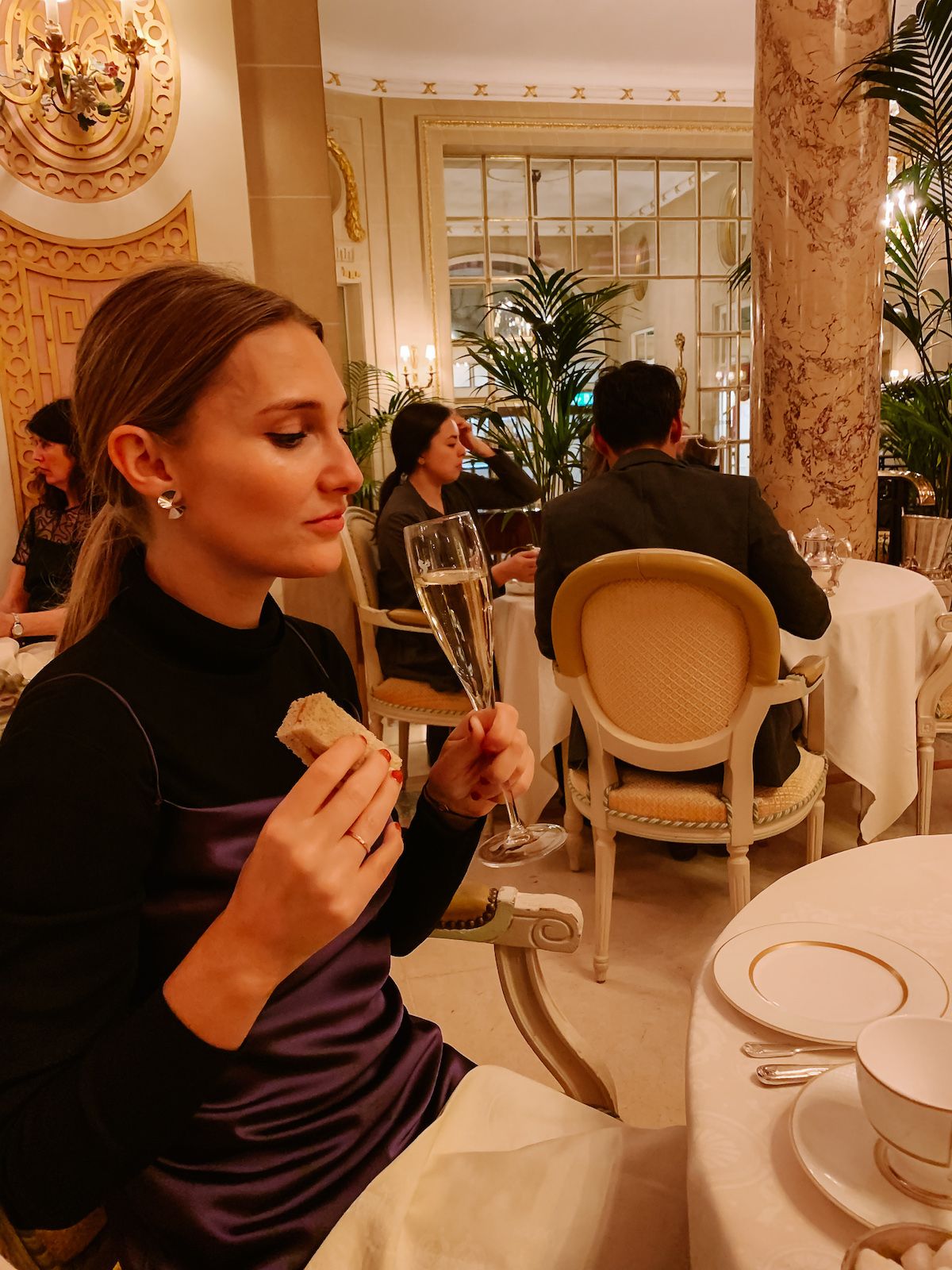 Cec in a purple satin dress, holding a glass of champagne and a tea sandwich in the luxurious tea room of The Ritz.