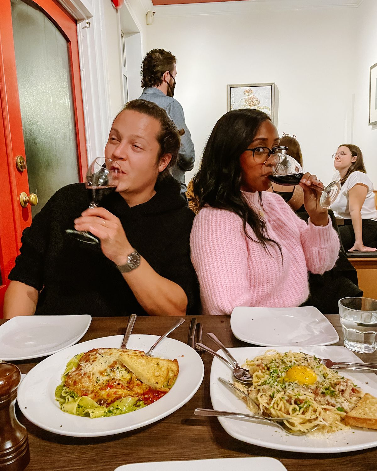 Ari and a friend enjoying red wine at Auntie Pesto's, with plates of fresh pasta and lasagna on a dark wood table.