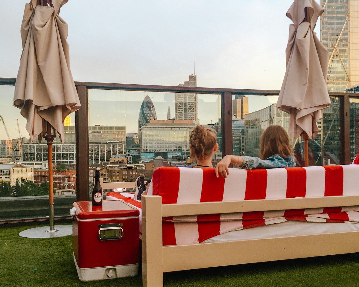 Cec and Katarina relaxing on a rooftop lounge with a red-and-white striped couch, enjoying a city view with The Gherkin.