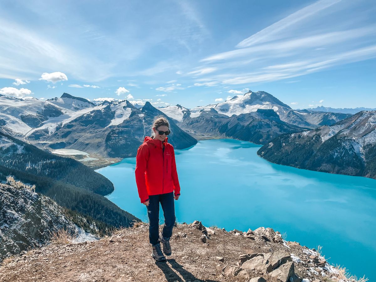 Cec in a red jacket stands on a rocky ledge overlooking the bright blue Garibaldi Lake and dramatic mountain ranges.