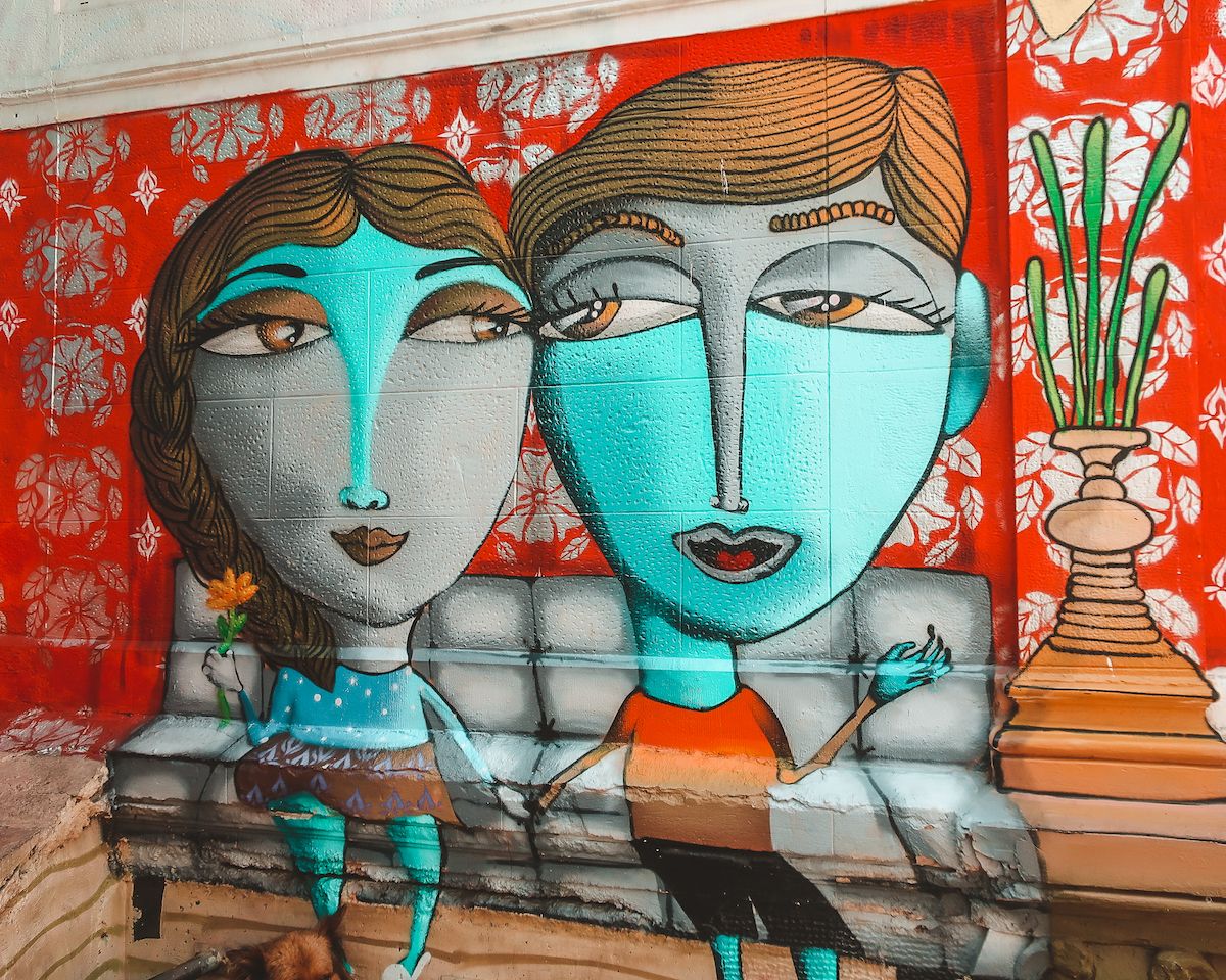  Mural of two blue-faced figures on a red wall in Valparaíso, with floral patterns and a couch illustration.