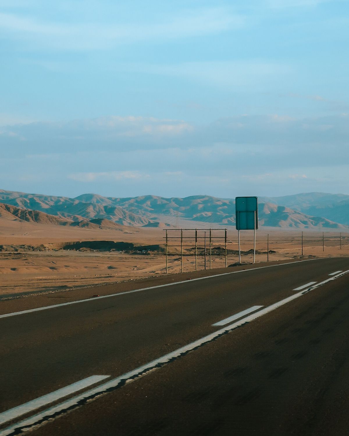 A quiet highway stretching across the arid Atacama Desert landscape, with rolling desert hills in the distance under a soft blue sky.