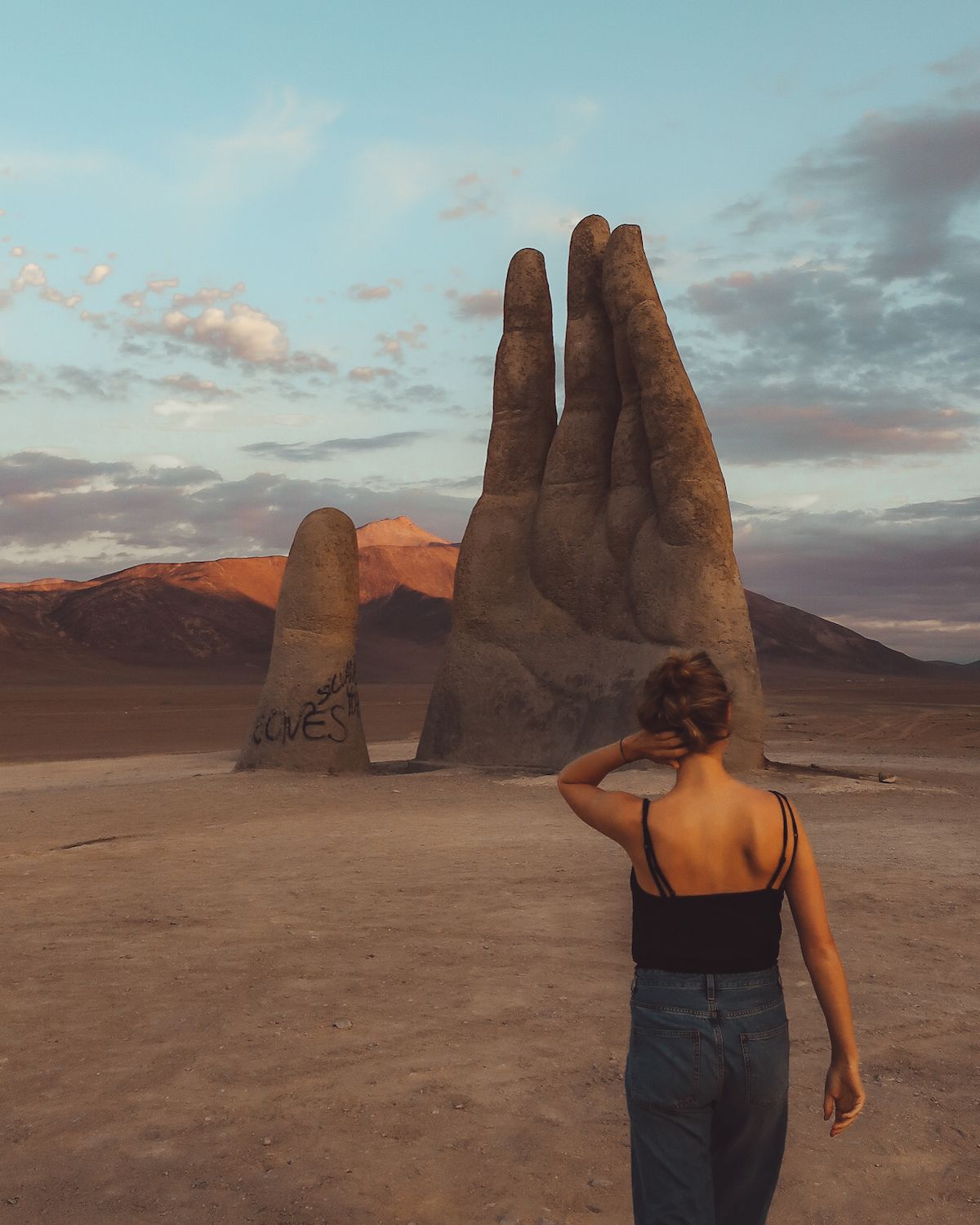 Cec walking towards the Hand of the Desert sculpture, a massive stone hand rising from the Atacama desert at sunset.