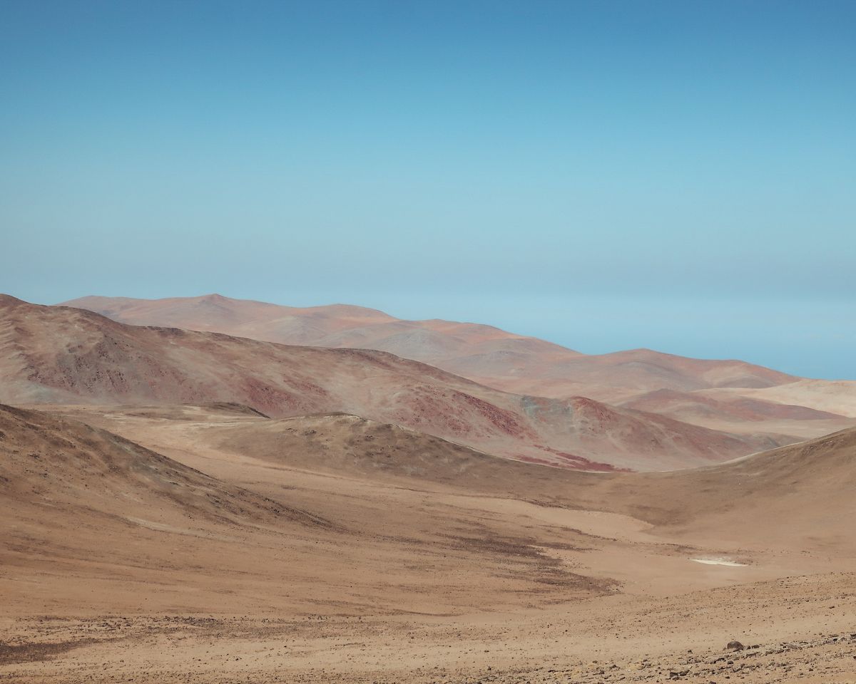 Rolling sand-coloured hills of Chile’s Atacama Desert stretching under a vast, pale blue sky with soft earthy hues.