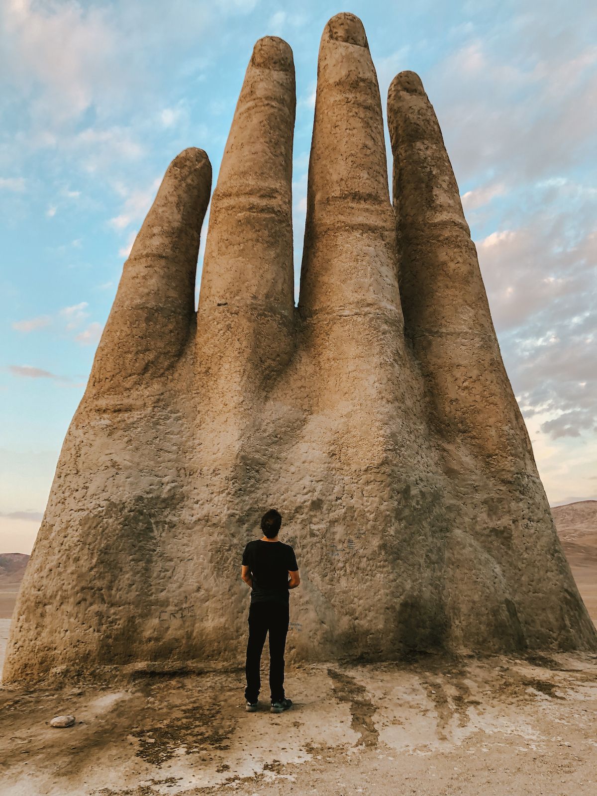 Ari standing in front of the Hand of the Desert, Chile, dwarfed by the towering stone fingers against a golden sky.