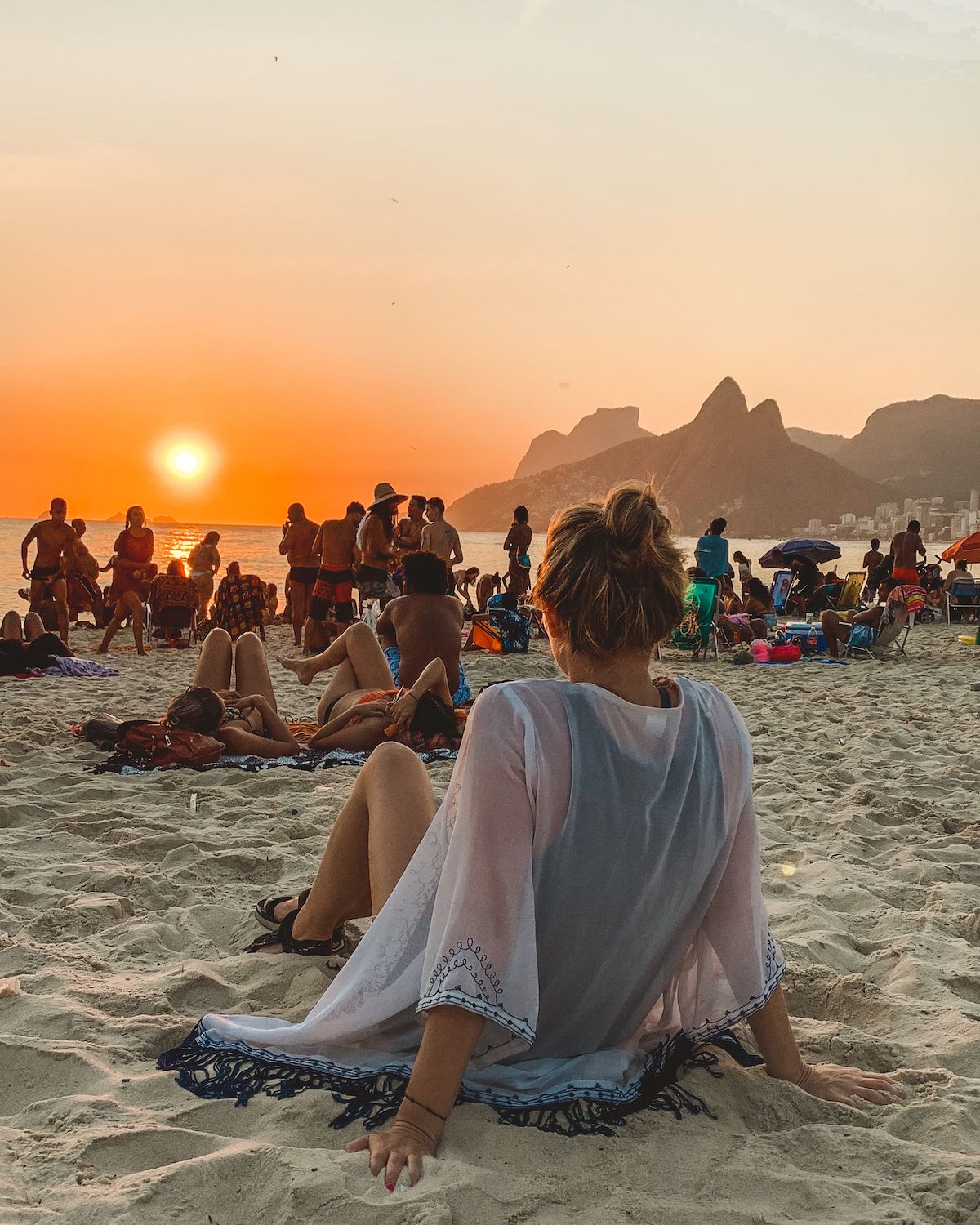 Cec sitting on Ipanema Beach at sunset, surrounded by people enjoying the golden hour with Two Brothers mountain in the distance.