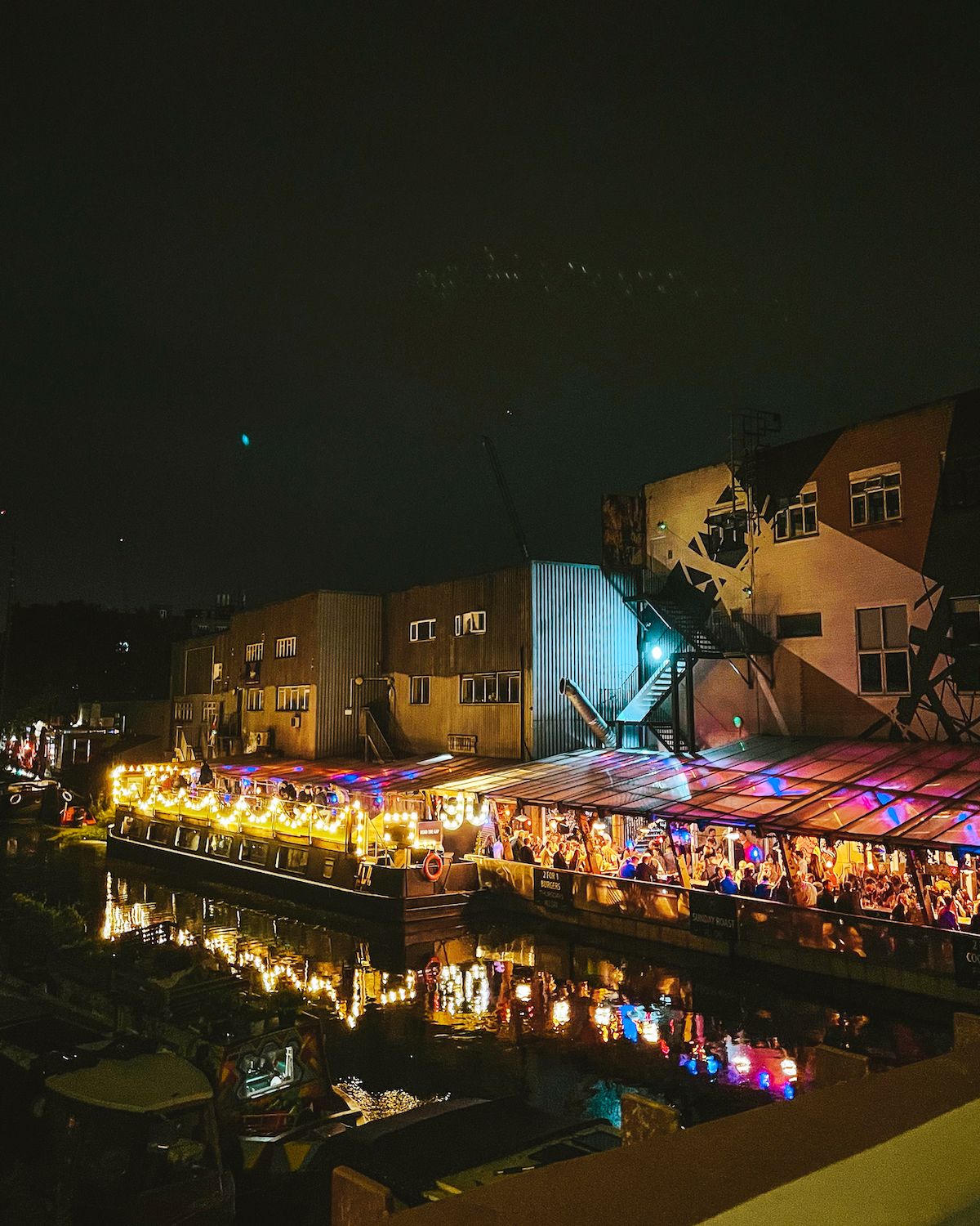 A lively canal-side bar in Hackney Wick glows with warm lights at night, reflecting on the water, with industrial buildings behind.