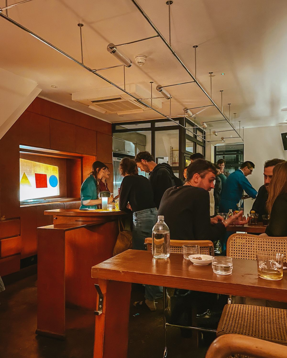 Stylish cocktail bar with a bauhaus decor, where patrons chat and sip drinks in a warmly lit, intimate space.