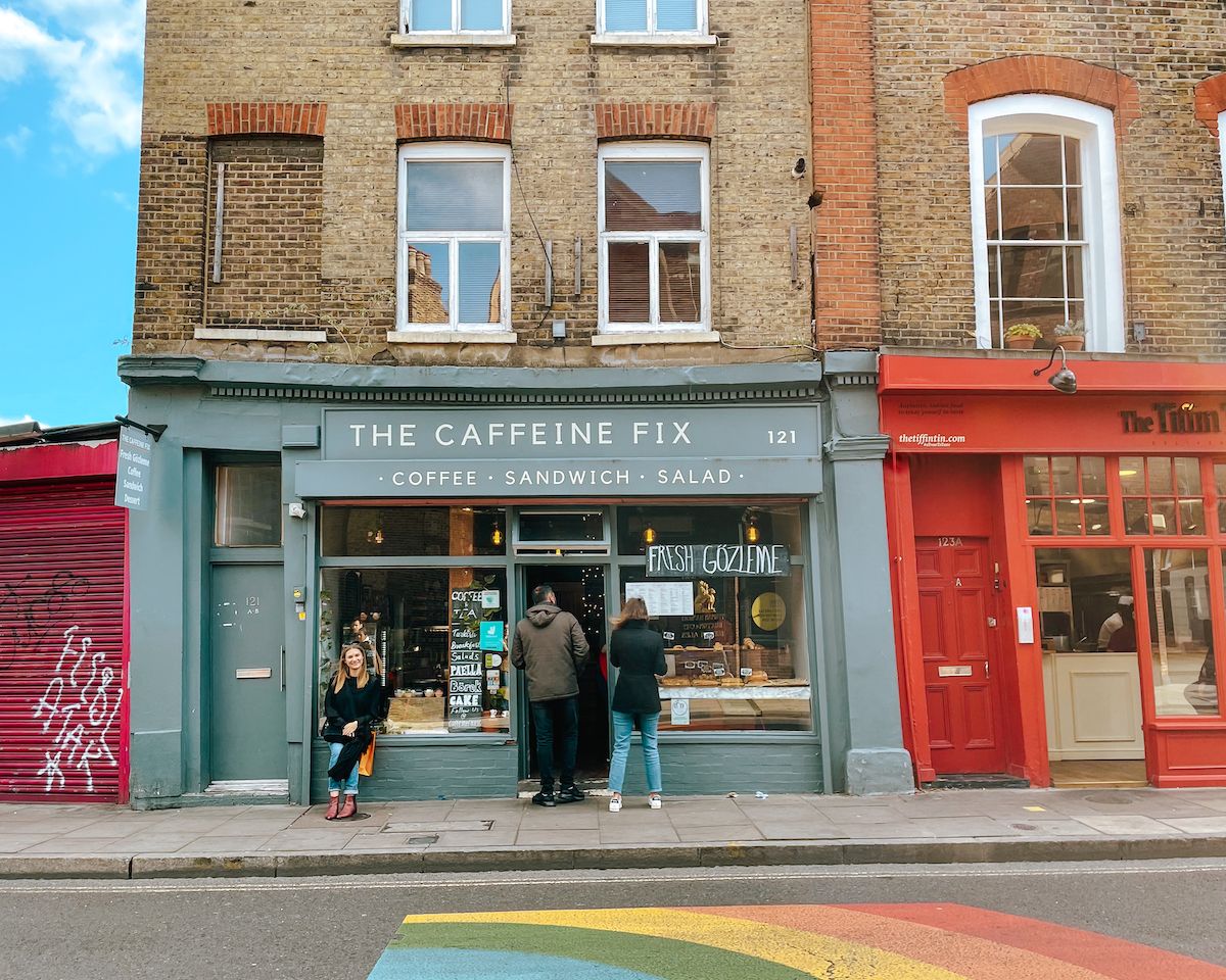 The Caffeine Fix cafe with a grey storefront and rainbow-painted sidewalk on a colourful Stoke Newington street.