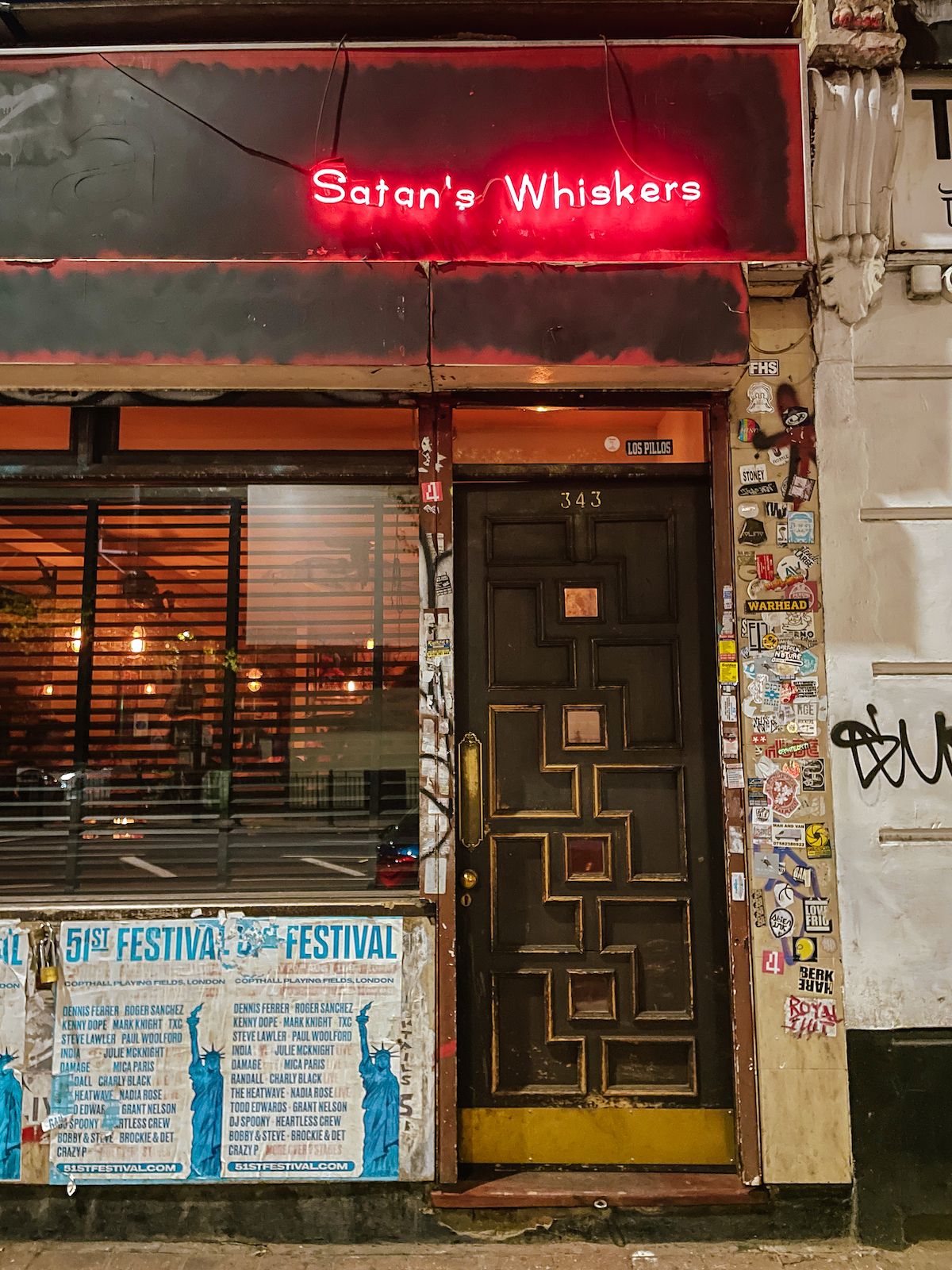 The entrance of Satan’s Whiskers, a moody East London cocktail bar with a neon red sign and a sticker-covered doorway.