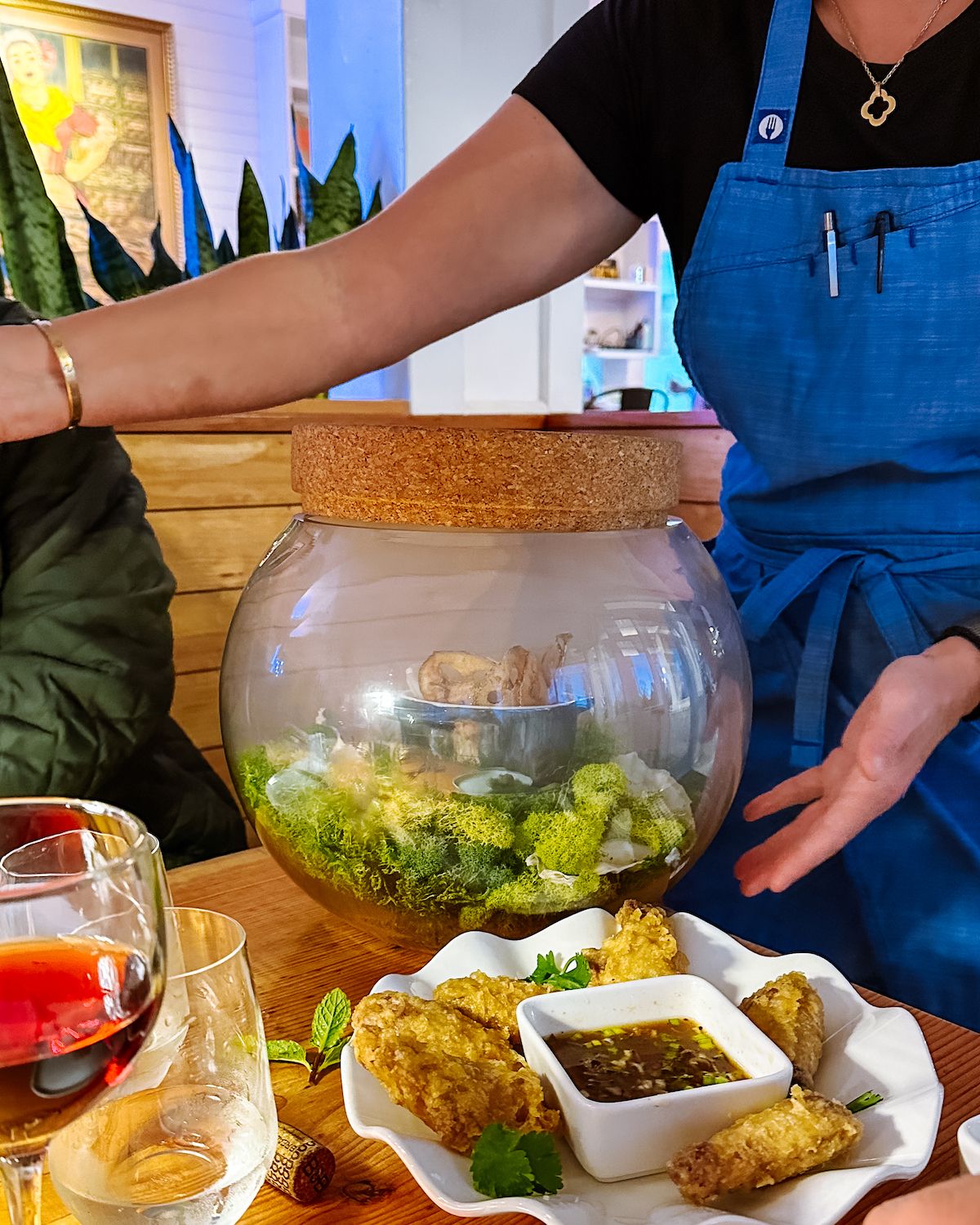 Server presenting a unique moss-lined glass bowl with food inside, alongside golden fried appetizers and dipping sauce at Off the Hook..