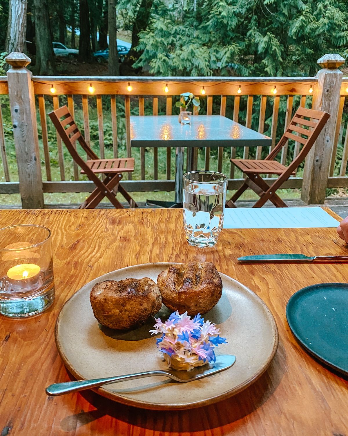 A wooden table set with popovers and butter covered in flowers, overlooking a forested patio with string lights.