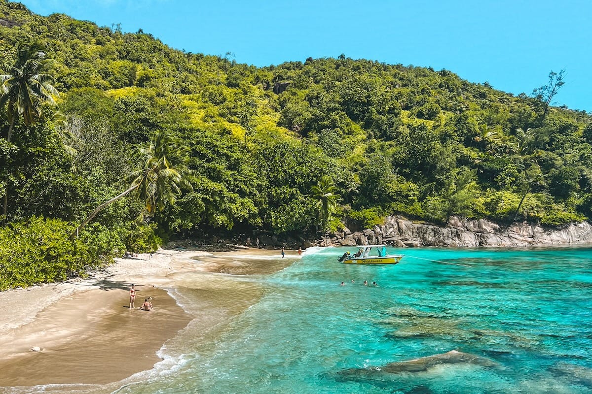 Snorkelers swim in turquoise waters near a sandy beach backed by lush green jungle and a small anchored boat in Seychelles.